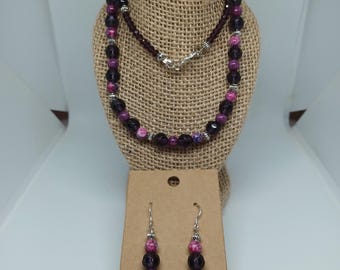Puede incluir: Un conjunto de collar y pendientes con cuentas moradas y rosas. El collar se exhibe en un busto de arpillera, con los pendientes colgados de una presentación de cartón. Las cuentas son una mezcla de formas redondas y facetadas, con detalles plateados.