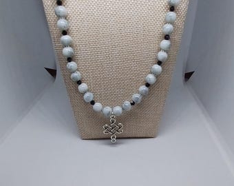 Puede incluir: Un collar de cuentas grises claras y negras, con un colgante de cruz celta plateado. El collar se exhibe sobre un busto de maniquí beige sobre un fondo blanco.