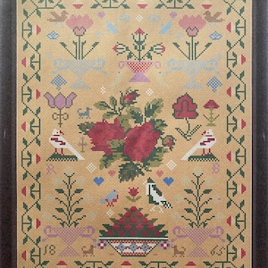 Puede incluir: Un sampler de punto de cruz enmarcado con un fondo beige. El diseño presenta rosas rojas, pájaros y motivos florales en rosa, azul y verde. El borde es un patrón geométrico repetitivo en verde y rojo. El número "18" es visible.