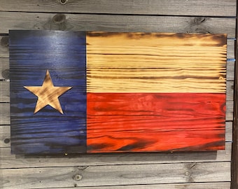 May include: A wooden Texas flag with a blue, white, and red color scheme. The flag is painted on a wooden plank and has a distressed look. The star is white and the stripes are red and white.