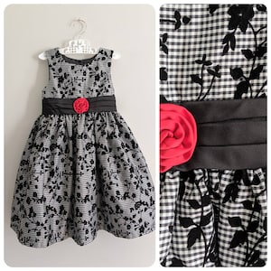 May include: A sleeveless dress with a black and white checkered pattern and black floral accents. The dress features a black satin sash and a red fabric flower at the waist. The dress is hanging on a white hanger.