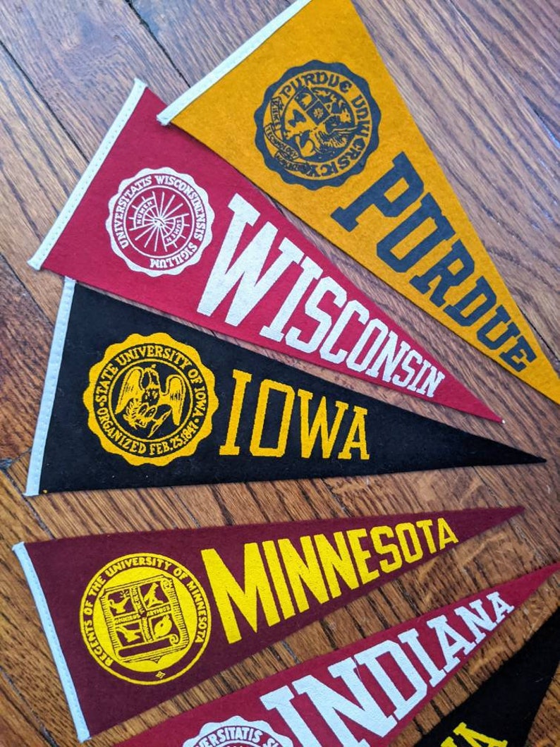 May include: A collection of felt pennants in various colors, each displaying a university name and emblem. The pennants include "Purdue," "Wisconsin," "Iowa," "Minnesota," and "Indiana," with white and yellow lettering and logos.