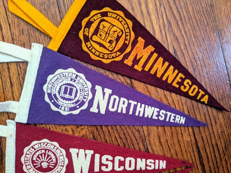 May include: Three vintage pennants in maroon, purple, and red. The maroon pennant reads "THE UNIVERSITY OF MINNESOTA". The purple pennant says "NORTHWESTERN". The red pennant says "WISCONSIN". Each pennant has a white border and a loop for hanging.