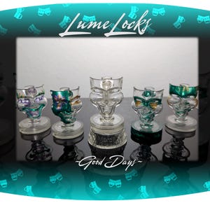 May include: Five glass pipes with different colored masks on the front. The pipes are on a black surface and the text "Lume Locks - Good Days -" is below them.