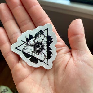 May include: A white sticker with a black floral design inside a black triangle. The sticker has a scalloped edge and is held in a hand.