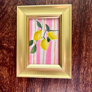 May include: A framed painting of lemons. The artwork features three yellow lemons on a branch with green leaves, set against a pink and white striped background. The frame is gold-colored.