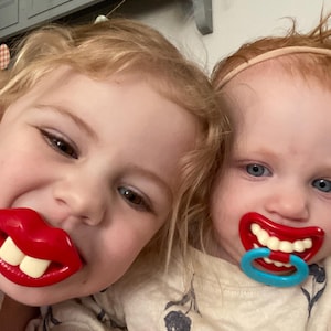 May include: Two young children smiling, each with a novelty pacifier shaped like a red mouth with white teeth. One pacifier has a blue ring. The children have blonde and red hair.