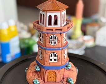 May include: A decorative lighthouse figurine in shades of orange, brown, and blue. The ceramic lighthouse features a detailed design with windows, a door, and a rooftop. Small blue and green floral accents are at the base of the lighthouse.