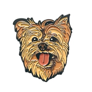 Puede incluir: Un pin de metal con la imagen de un Yorkshire Terrier. El pin tiene un contorno negro. El perro tiene pelo marrón y dorado y está sacando la lengua.
