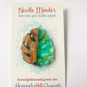 Puede incluir: Un protector de agujas con forma de hoja verde y marrón con purpurina dorada incrustada en la resina verde. El texto "Needle Minder" y "never lose your needles again!" está impreso en la tarjeta. El texto "heavenlyhellelements@msn.com" y "HeavenlyHell Elements" está impreso en la tarjeta.