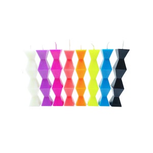 May include: A set of eight colorful candles, each with a unique geometric design. The candles are arranged in a row from white to black, with shades of purple, pink, orange, yellow, and blue in between.