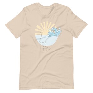 Puede incluir: Una camiseta beige con un gr&aacute;fico de una cara estilizada con un sol y olas en el ojo. El texto "Wild Waves" est&aacute; escrito en el gr&aacute;fico.
