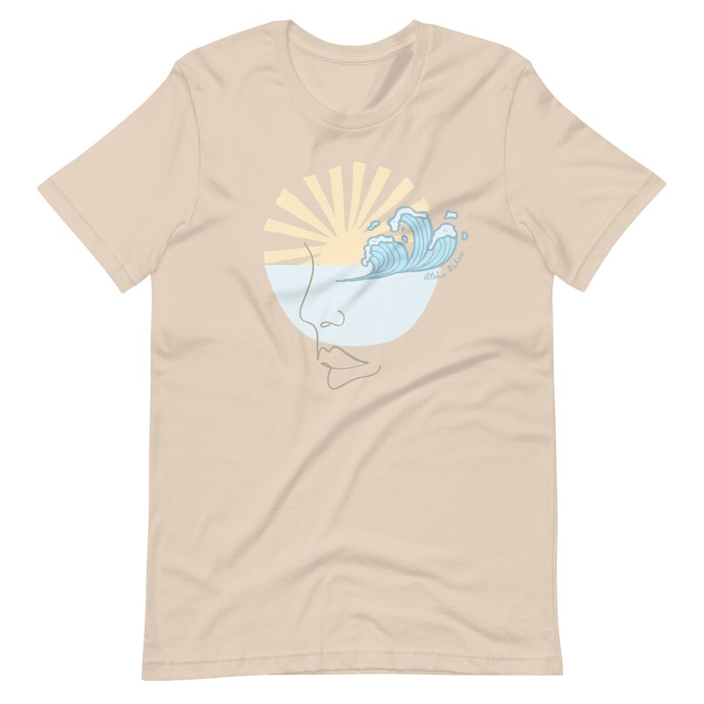 Puede incluir: Una camiseta beige con un gr&aacute;fico de una cara estilizada con un sol y olas en el ojo. El texto "Wild Waves" est&aacute; escrito en el gr&aacute;fico.