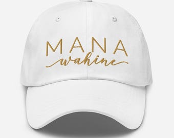 Puede incluir: Una gorra de béisbol blanca con el texto MANA wahine bordado en hilo dorado en la parte frontal. La palabra MANA está en una fuente sans-serif limpia, mientras que la palabra wahine está escrita en una fuente cursiva debajo de ella.