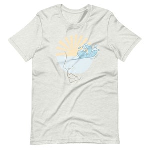 Puede incluir: Una camiseta gris claro con un dise&ntilde;o gr&aacute;fico de una cara estilizada con una ola rompiendo sobre el ojo. El sol se pone detr&aacute;s de la cara y el texto "Aloha Melon" est&aacute; escrito en la ola.