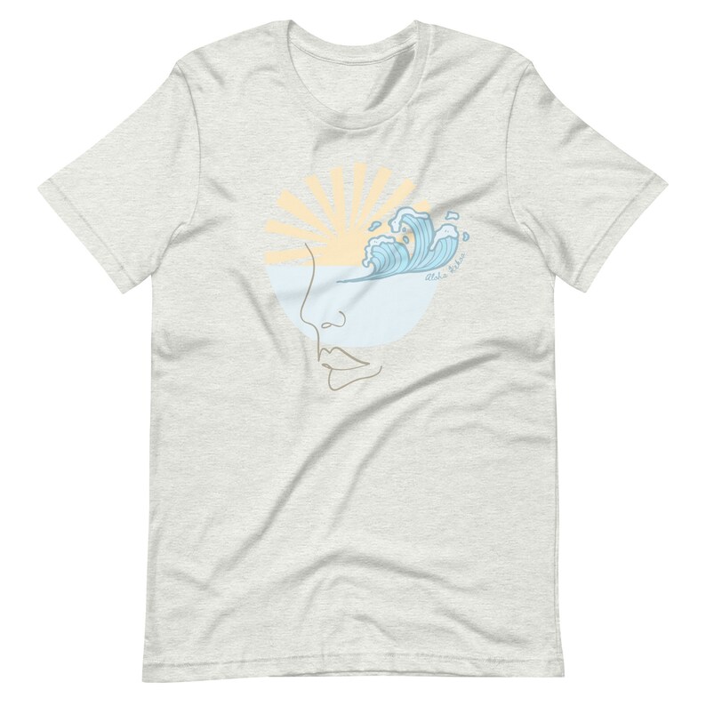 Puede incluir: Una camiseta gris claro con un dise&ntilde;o gr&aacute;fico de una cara estilizada con una ola rompiendo sobre el ojo. El sol se pone detr&aacute;s de la cara y el texto "Aloha Melon" est&aacute; escrito en la ola.