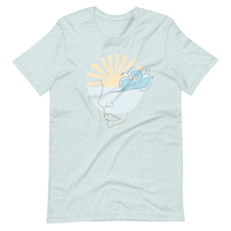 Puede incluir: Una camiseta azul claro con un gr&aacute;fico de una cara estilizada con una ola rompiendo sobre la parte superior de la cabeza. El sol brilla detr&aacute;s de la ola. El texto "Ride the Wave" est&aacute; escrito debajo de la ola.