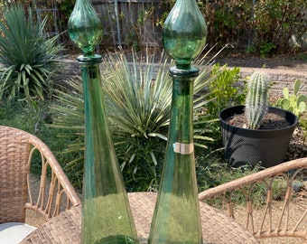 May include: Two tall, green glass bottles with elongated, teardrop-shaped stoppers. The bottles have a conical shape and are displayed on a woven table. The background features a garden setting with plants and a wooden fence.