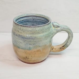May include: Handmade ceramic mug with a rounded body and a large handle. The mug features a color gradient, transitioning from a light blue interior to a light green and tan exterior. The mug is suitable for coffee, tea, or other beverages.
