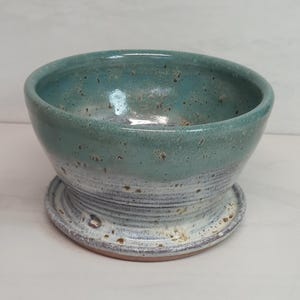 May include: A handmade ceramic bowl with a teal and white glaze. The bowl has a rounded shape and sits on a matching base. The exterior features a speckled texture, adding to its rustic charm. Ideal for serving or as a decorative piece.