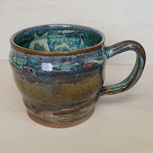 May include: A handmade ceramic mug with a rounded body and a large handle. The mug features a blend of blue, green, and brown glazes, creating a unique, textured appearance. The interior has a lighter blue and yellow glaze.