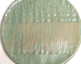 May include: A light green ceramic plate with a raised design of a row of trees with leaves on top. The plate is oval shaped.