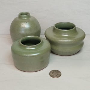 May include: Three handmade ceramic vases in varying sizes and shapes. The vases are a muted green color with a smooth, glossy finish. The smallest vase is a low, rounded cylinder, while the largest is a bulbous shape. A quarter coin is next to the vases.
