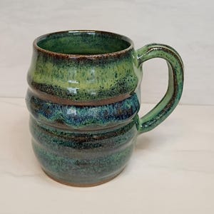 May include: Handmade ceramic mug with a unique, textured design. The mug features a blend of green and blue hues, with a brown band around the middle. The handle is a matching green, and the mug is suitable for hot or cold beverages.