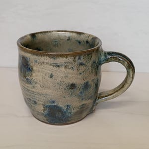 May include: A handmade ceramic mug with a rounded body and a sturdy handle. The mug features a speckled, textured surface with shades of beige, brown, and blue. The interior and rim are a darker brown. A unique piece for enjoying coffee or tea.