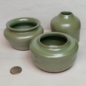 May include: Three handmade, sage green ceramic vessels of varying shapes. The vessels include a small, round vase, a low, wide bowl, and a slightly taller, rounded pot. A US quarter is next to the vessels for scale.