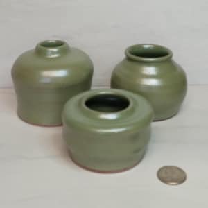 May include: Three sage green ceramic vases of varying sizes. The vases have a rounded shape with a small opening at the top. The smallest vase is a low cylinder. The largest vase is a rounded shape. The vases are on a white surface.