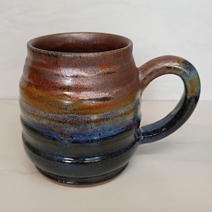 May include: A handmade ceramic mug with a unique, layered color scheme. The mug transitions from a dark brown rim to bands of orange, blue, and black. The handle is a dark blue, complementing the base. The mug has a textured, ridged design.