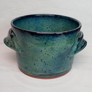 May include: A handmade ceramic pot with a teal and green glaze. The pot has two handles and a reddish-brown base. The interior and rim are a darker shade of teal. The pot is a unique piece of pottery.