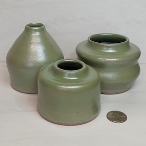 May include: Three handmade, sage green ceramic vases of varying shapes and sizes. The vases have a matte finish and a slightly speckled texture. The smallest vase is cylindrical, the middle one is rounded, and the largest is bulbous. A US quarter is next to the vases.