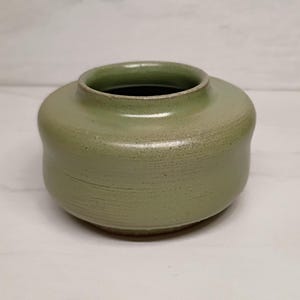 May include: A handcrafted, sage green ceramic vase with a rounded body and a wide opening. The vase has a textured surface and a dark interior. The design is simple and elegant, suitable for displaying flowers or as a decorative piece.