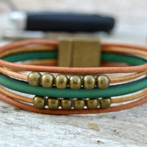 May include: A multi-strand bracelet featuring brown, green, and tan leather cords. The bracelet is accented with brass-colored beads and a square clasp. The design is simple and stylish, suitable for everyday wear.