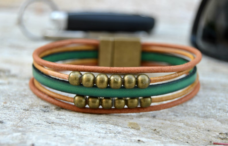 May include: A multi-strand bracelet featuring brown, green, and tan leather cords. The bracelet is accented with brass-colored beads and a square clasp. The design is simple and stylish, suitable for everyday wear.