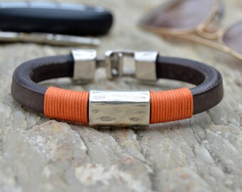 May include: A brown leather bracelet with an orange cord wrap and a silver metal clasp. The bracelet has a silver metal bar with a hammered texture.