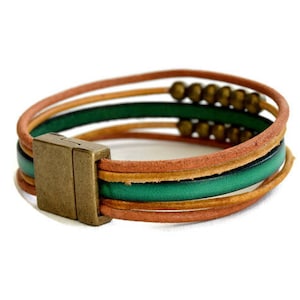 May include: A multi-strand leather bracelet with a magnetic clasp. The bracelet features layers of brown, green, and tan leather strands. Small brass beads are strung along one of the strands. The clasp is a rectangular shape with a bronze finish.