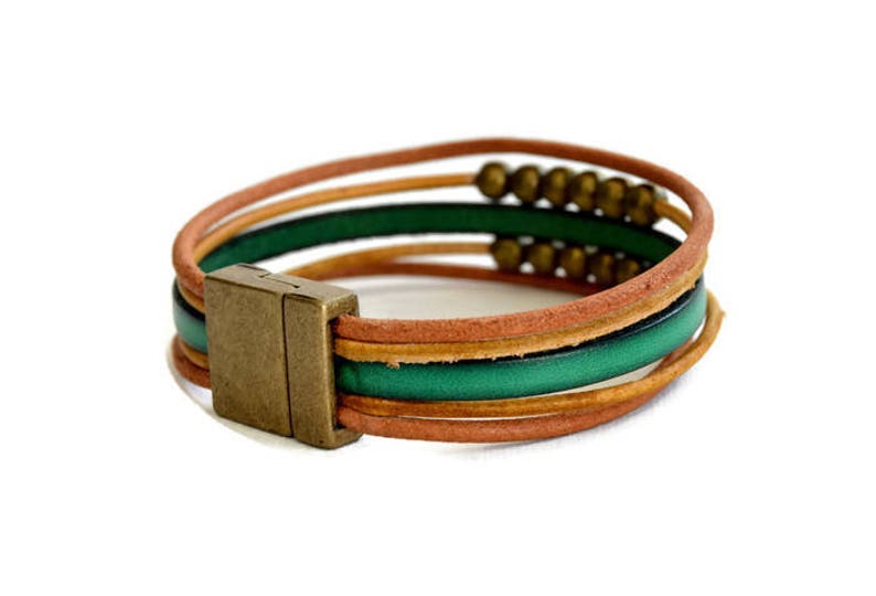 May include: A multi-strand leather bracelet with a magnetic clasp. The bracelet features layers of brown, green, and tan leather strands. Small brass beads are strung along one of the strands. The clasp is a rectangular shape with a bronze finish.