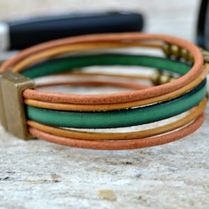 May include: Leather bracelet with a magnetic clasp. The bracelet features multiple strands in shades of brown and green, accented with small gold-colored beads. The clasp is a rectangular shape in a bronze tone.