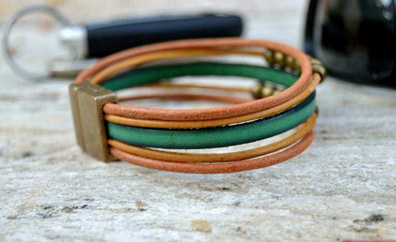 May include: Leather bracelet with a magnetic clasp. The bracelet features multiple strands in shades of brown and green, accented with small gold-colored beads. The clasp is a rectangular shape in a bronze tone.