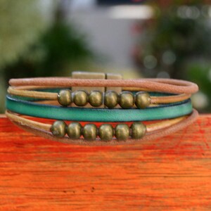 May include: A multi-strand bracelet featuring a combination of brown, green, and tan leather cords. The bracelet is adorned with small, round, bronze-colored beads and a bronze-tone clasp. The design is casual and bohemian.