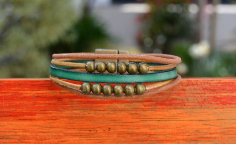 May include: A multi-strand bracelet featuring a combination of brown, green, and tan leather cords. The bracelet is adorned with small, round, bronze-colored beads and a bronze-tone clasp. The design is casual and bohemian.