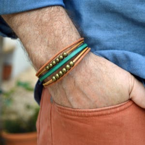 May include: A multi-strand bracelet with brown, green, and gold accents. The bracelet features a tan leather band, a vibrant green band, and is adorned with small, gold-colored beads. The bracelet is worn on the wrist.