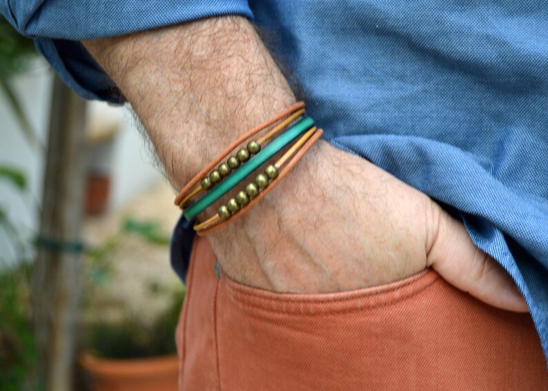 May include: A multi-strand bracelet with brown, green, and gold accents. The bracelet features a tan leather band, a vibrant green band, and is adorned with small, gold-colored beads. The bracelet is worn on the wrist.