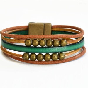May include: A multi-strand bracelet featuring brown, green, and tan leather cords. The bracelet is accented with gold-colored beads and a rectangular clasp. The design is simple and elegant, suitable for everyday wear.