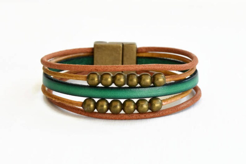May include: A multi-strand bracelet featuring brown, green, and tan leather cords. The bracelet is accented with gold-colored beads and a rectangular clasp. The design is simple and elegant, suitable for everyday wear.