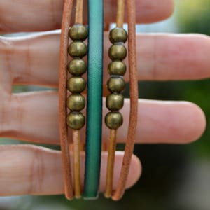 May include: A multi-strand bracelet featuring tan leather cords, a teal leather band, and rows of bronze-colored beads. The bracelet is designed with a casual, bohemian style, suitable for everyday wear. The bracelet is approximately 0.5 inches wide.
