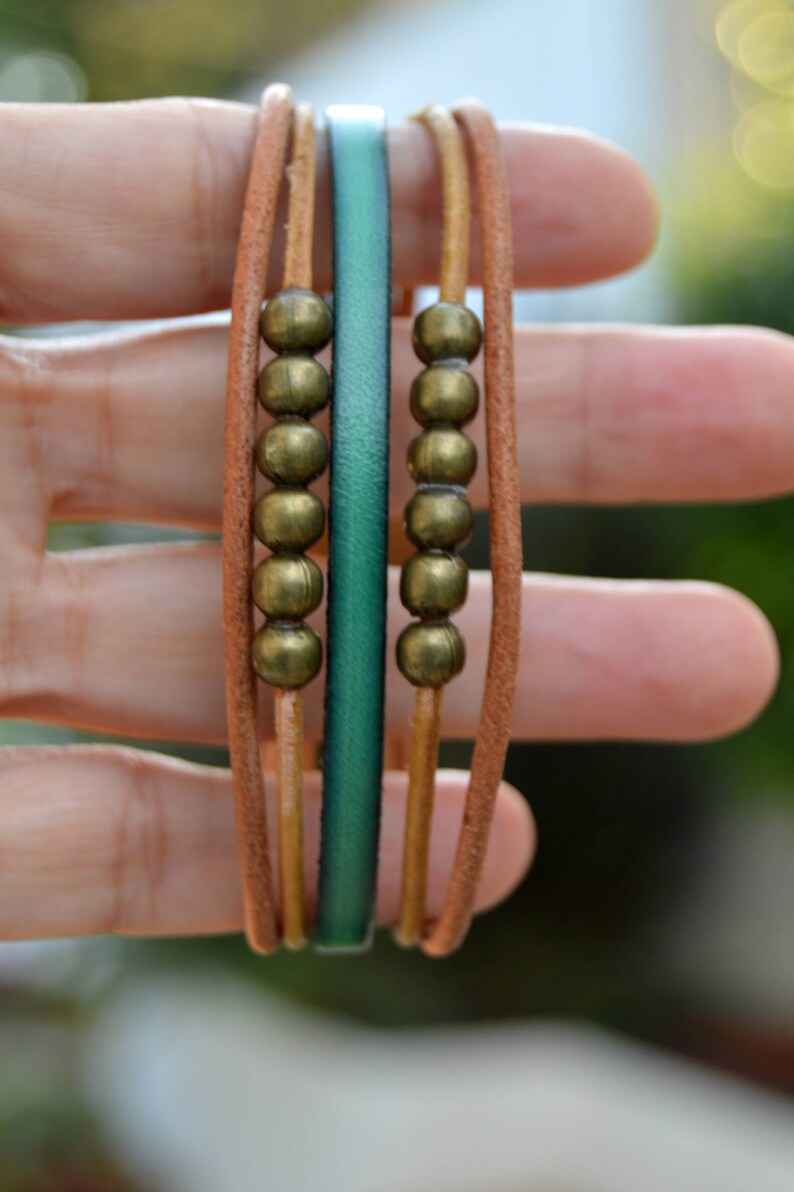 May include: A multi-strand bracelet featuring tan leather cords, a teal leather band, and rows of bronze-colored beads. The bracelet is designed with a casual, bohemian style, suitable for everyday wear. The bracelet is approximately 0.5 inches wide.